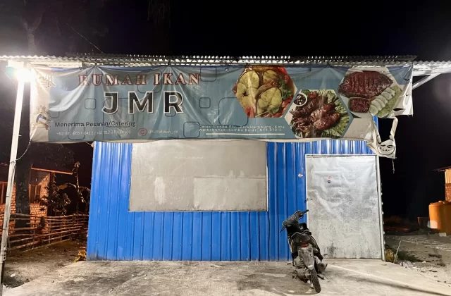 The exterior of Rumah Ikan JMR, a blue corrugated metal building at night, features a banner displaying various fish dishes above a parked motorcycle.