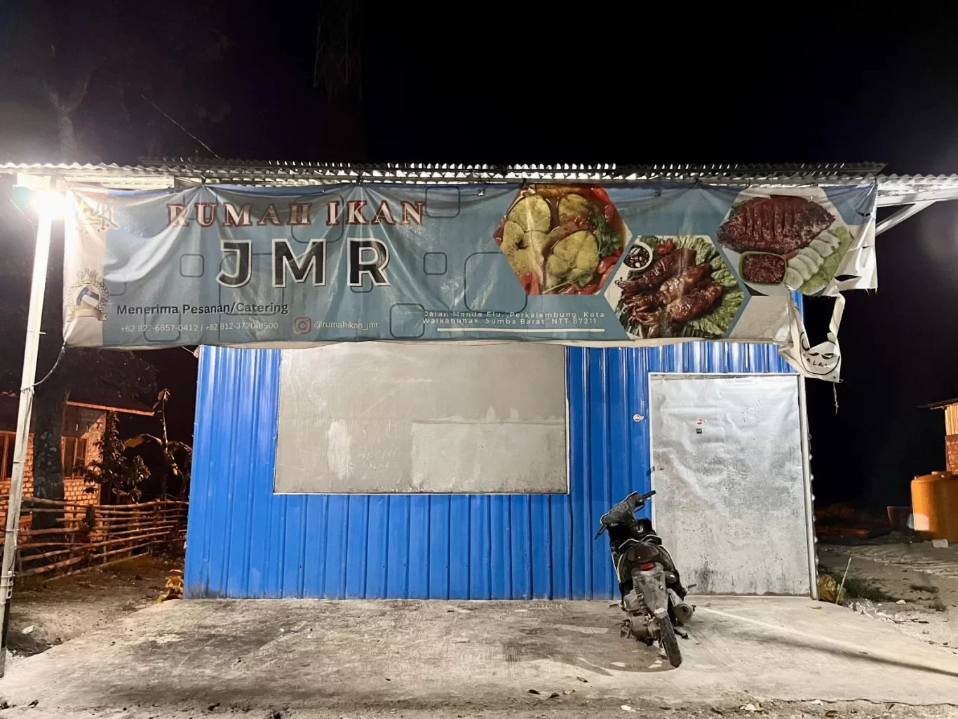 The exterior of Rumah Ikan JMR, a blue corrugated metal building at night, features a banner displaying various fish dishes above a parked motorcycle.