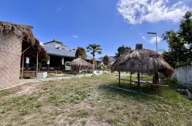 Traditional thatched-roof dining huts and a main building with a green roof are visible under a bright blue sky at Marapu Umma in West Sumba.