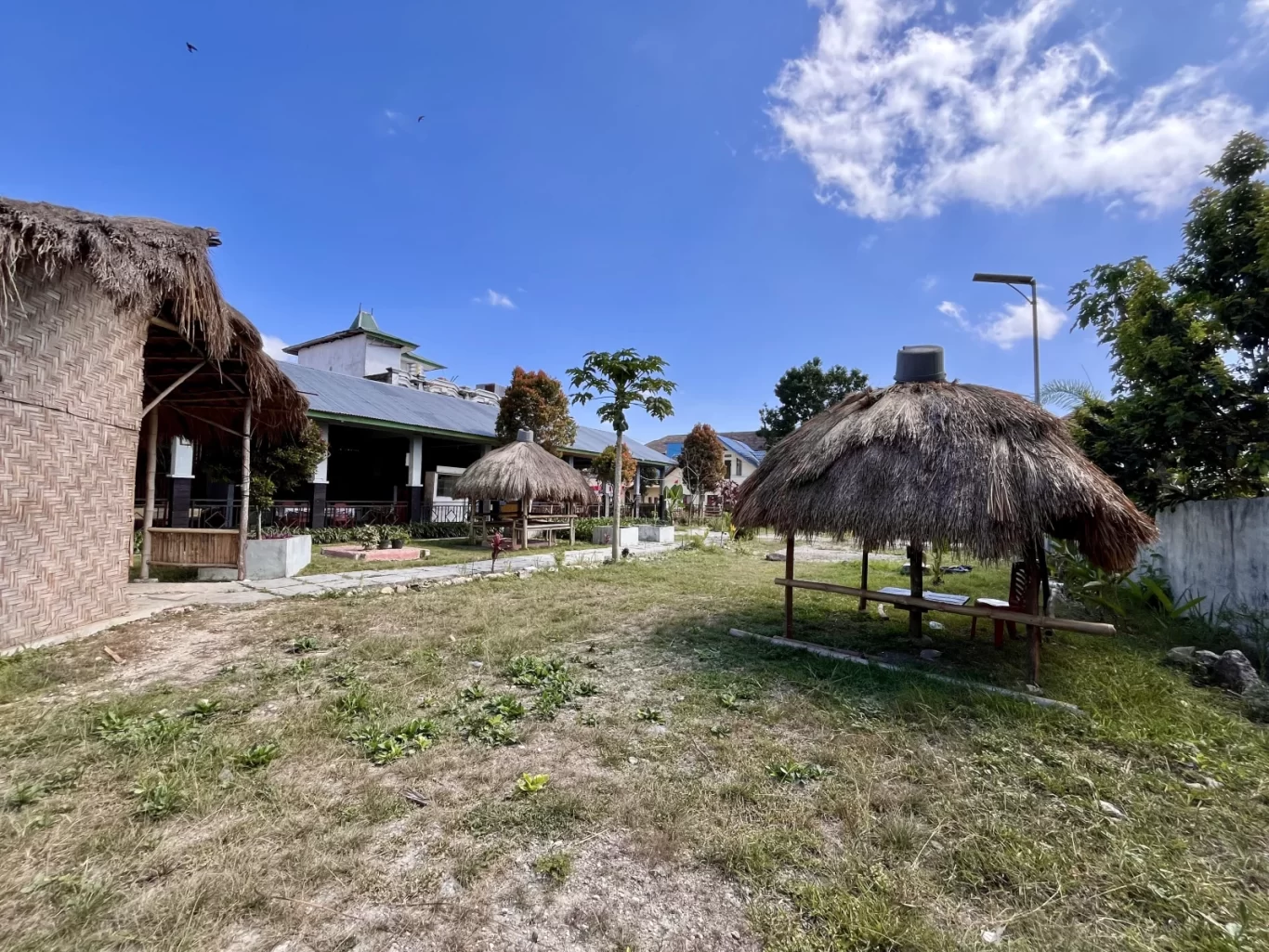 Traditional thatched-roof dining huts and a main building with a green roof are visible under a bright blue sky at Marapu Umma in West Sumba.
