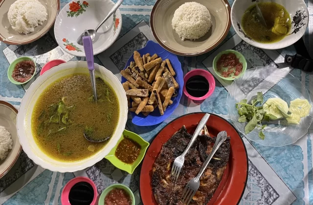 A vibrant overhead view of an Indonesian seafood feast, including grilled fish, soup, rice, fried items, and various sauces, laid out on a blue patterned table.