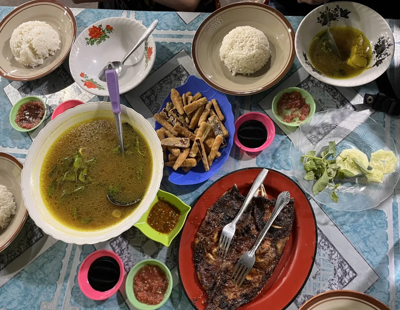 A vibrant overhead view of an Indonesian seafood feast, including grilled fish, soup, rice, fried items, and various sauces, laid out on a blue patterned table.