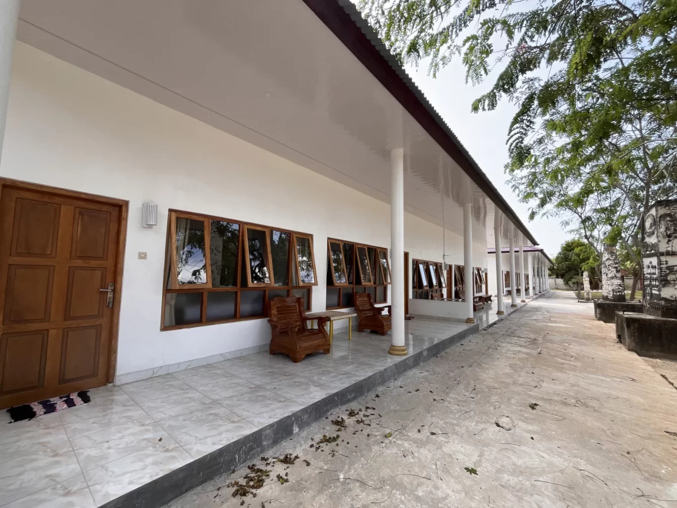 A long, white hotel building with a covered, tiled walkway, brown wooden doors and windows, next to a path with trees and stone monuments.