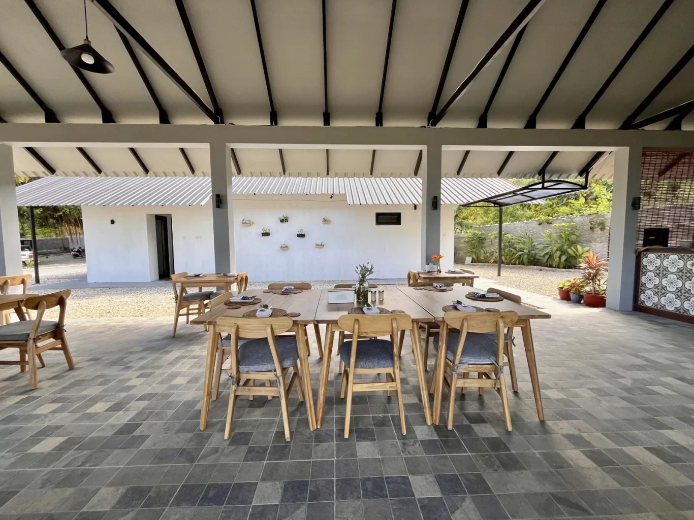 A spacious open-air restaurant dining area features wooden tables and chairs on a dark tiled floor, with white buildings and lush greenery in the background.