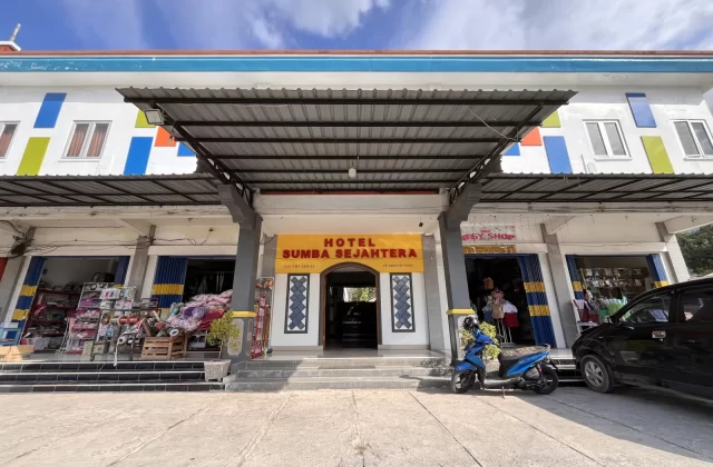 The colorful Sumba Sejahtera Hotel entrance in Tambolaka, Indonesia, features a yellow banner, shops, a blue scooter, and a black car.