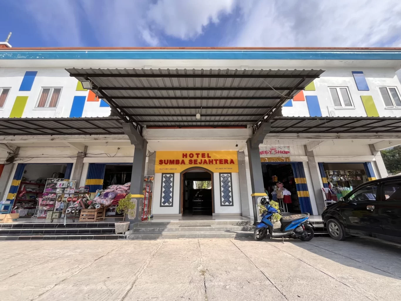 The colorful Sumba Sejahtera Hotel entrance in Tambolaka, Indonesia, features a yellow banner, shops, a blue scooter, and a black car.