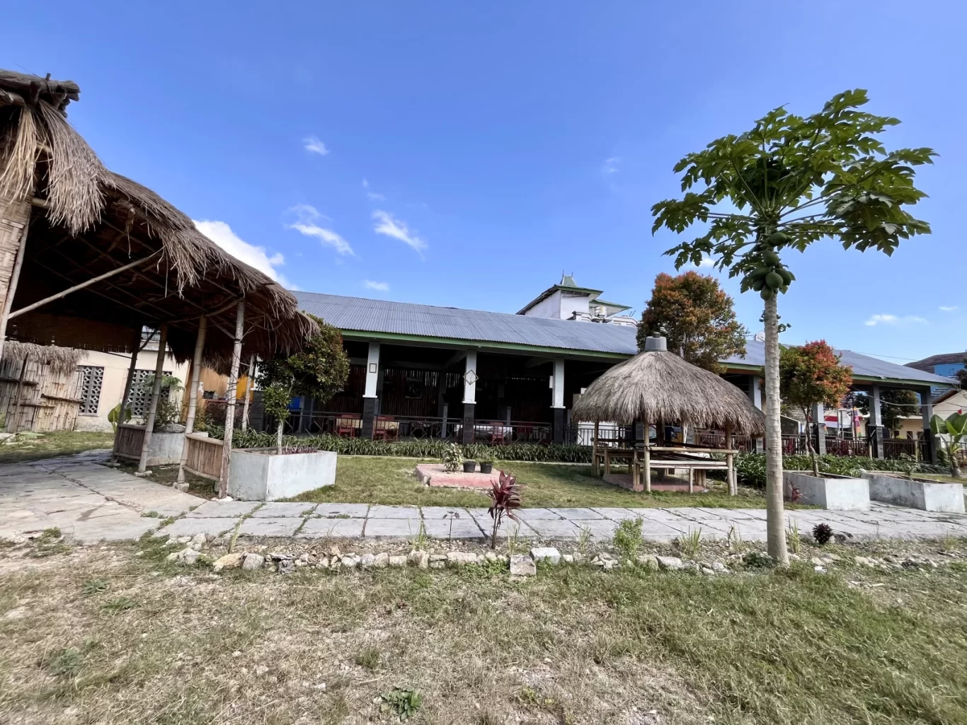 Marapu Umma restaurant features traditional thatched-roof pavilions, a large building, a papaya tree, and green grass under a clear blue sky.