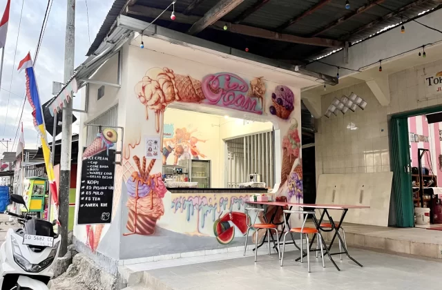 A small white ice cream shop with vibrant murals of melting ice cream cones and sundaes on its exterior walls. Two red tables with orange chairs sit outside, and an Indonesian flag flies nearby under a cloudy sky.
