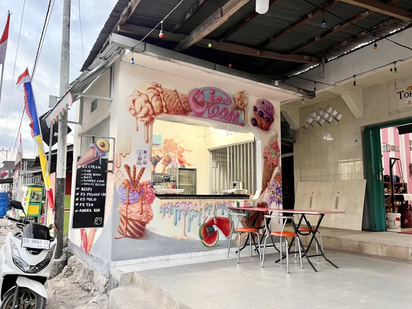 A small white ice cream shop with vibrant murals of melting ice cream cones and sundaes on its exterior walls. Two red tables with orange chairs sit outside, and an Indonesian flag flies nearby under a cloudy sky.