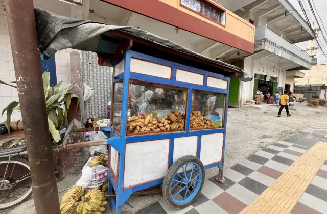 A blue and white food cart, laden with golden-brown Indonesian fritters and a pile of yellow bananas, sits on a tiled sidewalk in front of a building.
