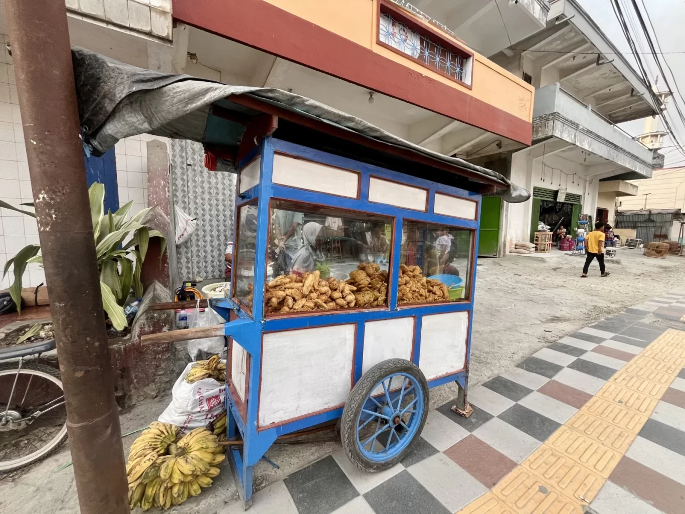 A blue and white food cart, laden with golden-brown Indonesian fritters and a pile of yellow bananas, sits on a tiled sidewalk in front of a building.