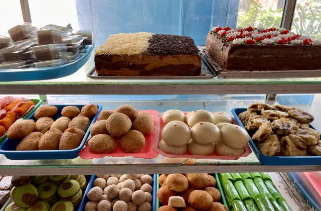 A glass display case filled with various Indonesian traditional snacks and cakes, including decorated cakes, fried pastries, and green rolled treats.
