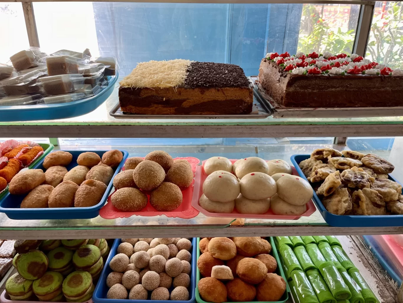 A glass display case filled with various Indonesian traditional snacks and cakes, including decorated cakes, fried pastries, and green rolled treats.