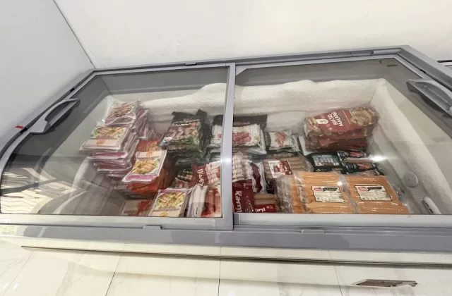 A large grey chest freezer with a glass lid is filled with various packaged frozen foods, including sausages and meatballs, set against a white tiled floor.