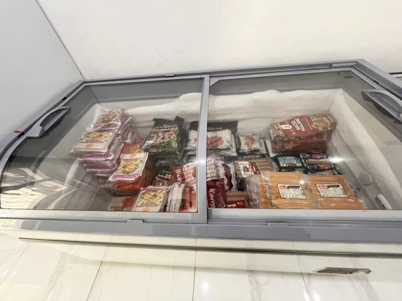 A large grey chest freezer with a glass lid is filled with various packaged frozen foods, including sausages and meatballs, set against a white tiled floor.