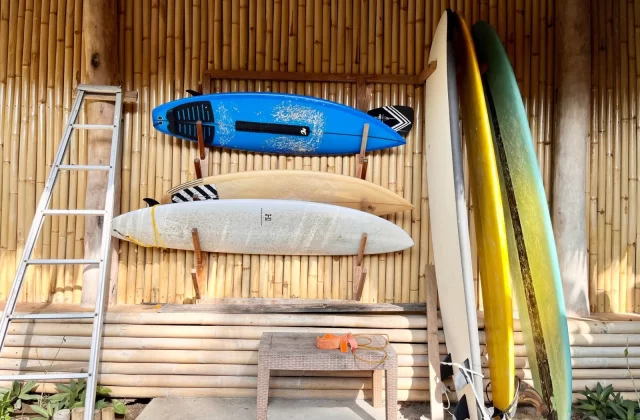 Several colorful surfboards, including blue, beige, white, yellow, and green, are stored against a natural bamboo wall next to a silver ladder and a small table.