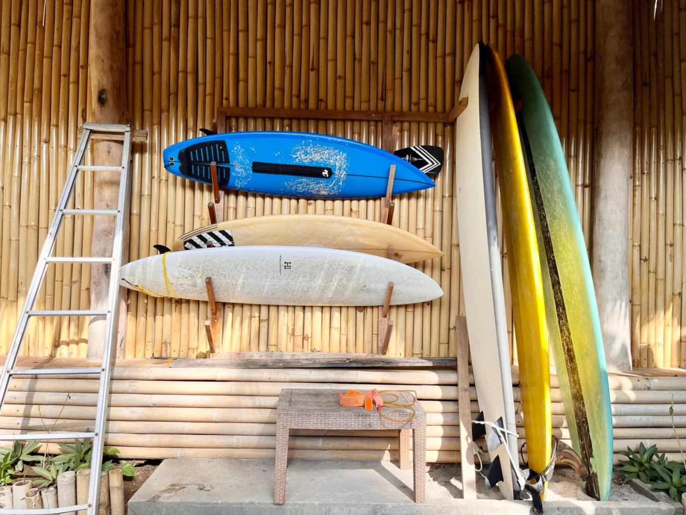 Several colorful surfboards, including blue, beige, white, yellow, and green, are stored against a natural bamboo wall next to a silver ladder and a small table.