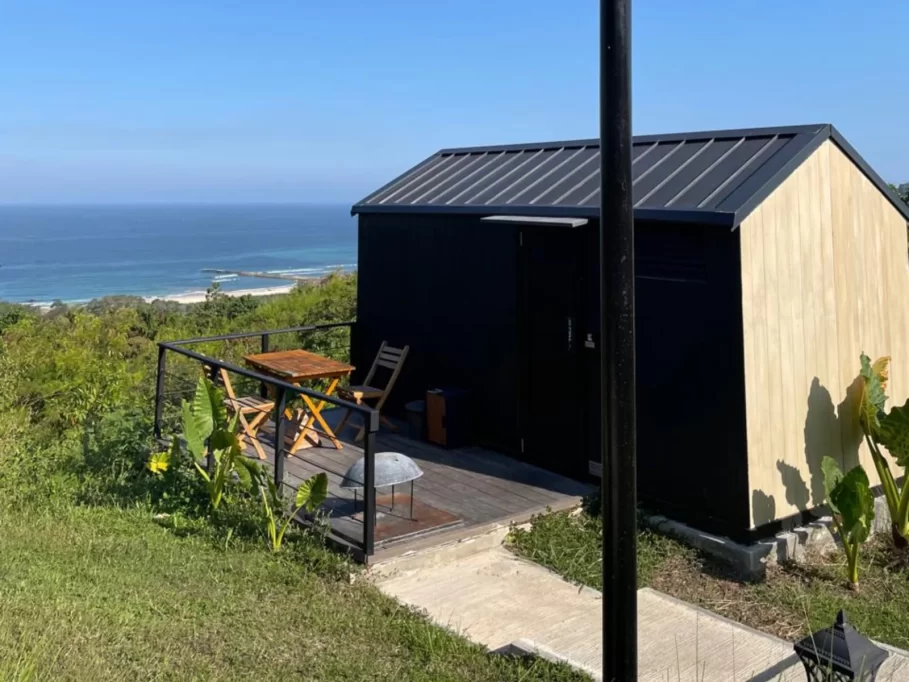 A black and light wood cabin with a deck and outdoor seating overlooks a lush green hillside, a white sand beach, and the clear blue ocean under a bright sky.