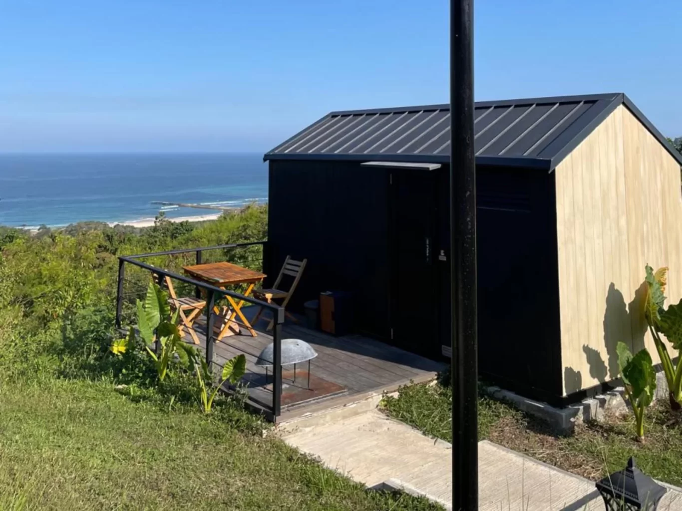 A black and light wood cabin with a deck and outdoor seating overlooks a lush green hillside, a white sand beach, and the clear blue ocean under a bright sky.