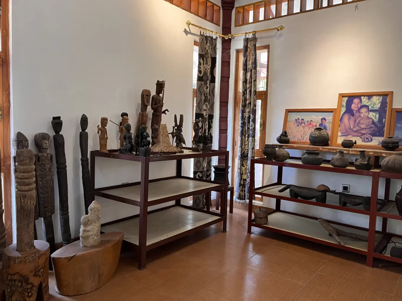 A bright museum room displays wooden carvings and dark pottery on brown shelves, with framed photographs on a white wall and patterned curtains by windows.
