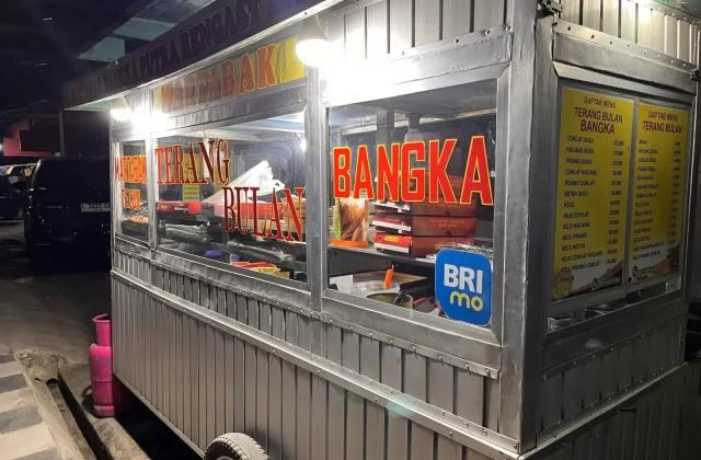 A brightly lit metal food stall, Martabak Bangka Putra Rengast, stands at night, displaying "Terang Bulan" and "Bangka" in red letters, with menu boards and a pink gas cylinder nearby.