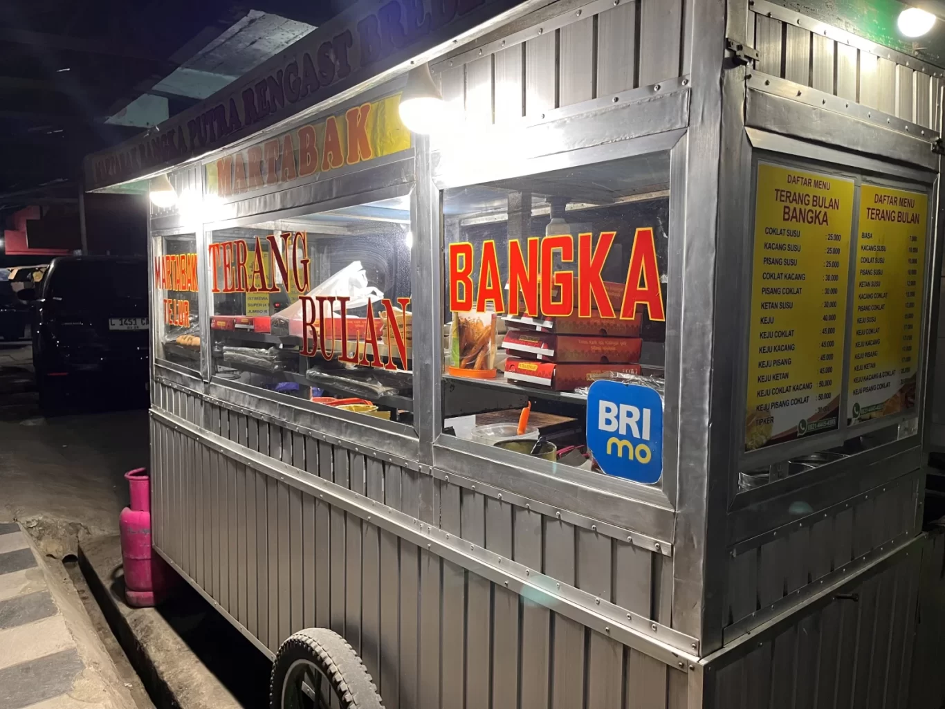 A brightly lit metal food stall, Martabak Bangka Putra Rengast, stands at night, displaying "Terang Bulan" and "Bangka" in red letters, with menu boards and a pink gas cylinder nearby.