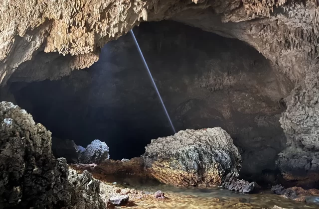 A beam of light shines from an opening in the ceiling of a dark, expansive cave, illuminating a clear water pool and textured rock formations.