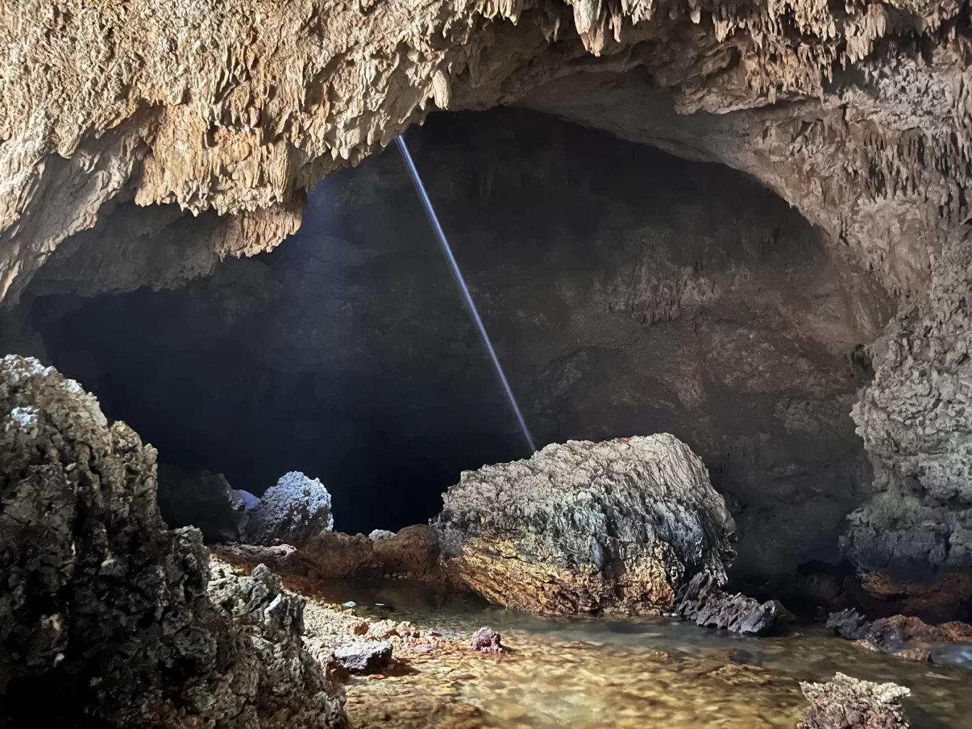 A beam of light shines from an opening in the ceiling of a dark, expansive cave, illuminating a clear water pool and textured rock formations.
