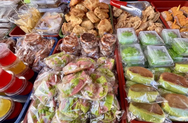 A street food stall displays numerous traditional Indonesian snacks, including golden-brown fried items, colorful layered cakes, and vibrant jelly drinks.