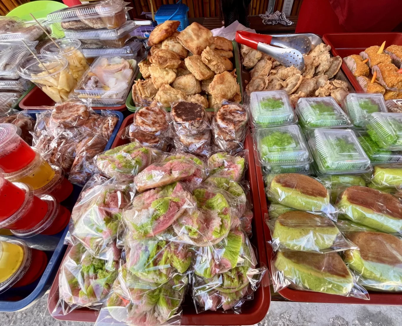 A street food stall displays numerous traditional Indonesian snacks, including golden-brown fried items, colorful layered cakes, and vibrant jelly drinks.