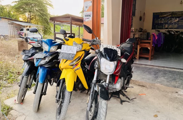 Several used motorcycles, including yellow, blue, black, and red models, are parked outside a shop in Tambolaka, Sumba.