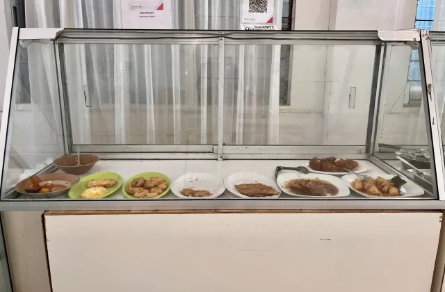 A glass display case holds multiple plates of Indonesian food, including fried items and sauced dishes, in a simple eatery with white curtains.