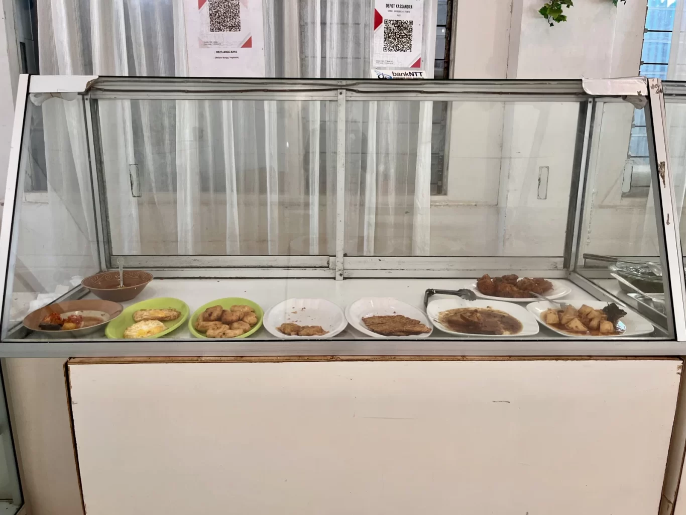 A glass display case holds multiple plates of Indonesian food, including fried items and sauced dishes, in a simple eatery with white curtains.