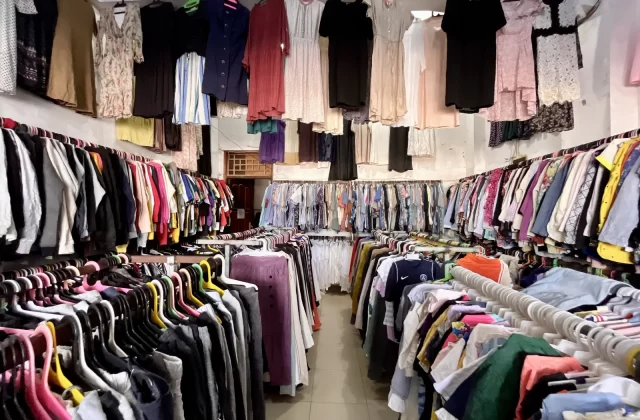 A brightly lit thrift store interior features long racks packed with diverse, colorful clothing for men, women, and children, with dresses hanging above.
