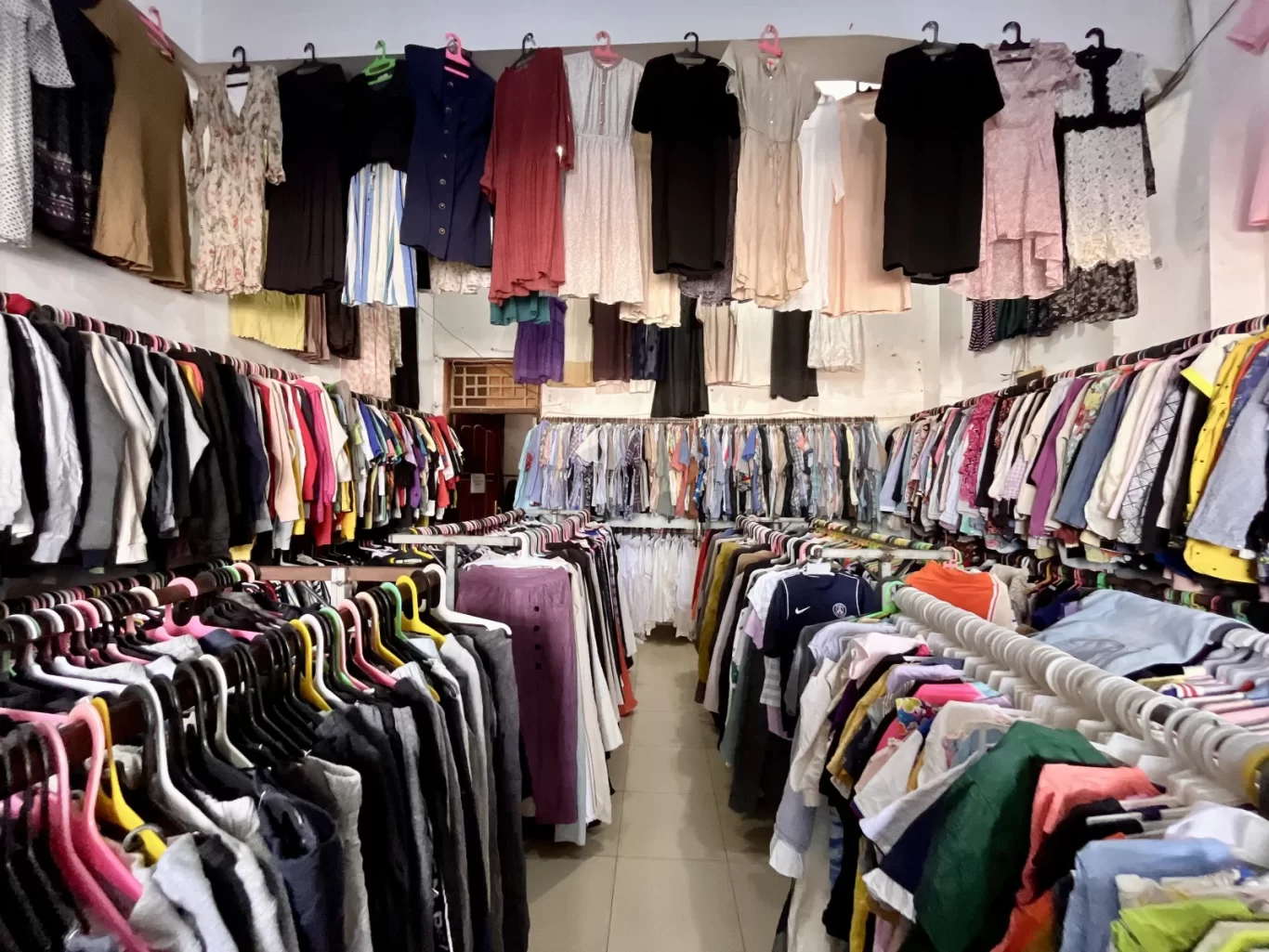 A brightly lit thrift store interior features long racks packed with diverse, colorful clothing for men, women, and children, with dresses hanging above.