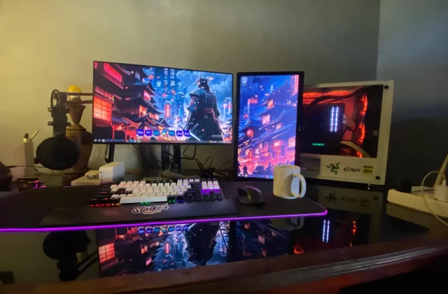 A dual monitor computer setup sits on a reflective dark desk, displaying vibrant blue and red cityscapes, with a white RGB keyboard and a gaming PC tower.