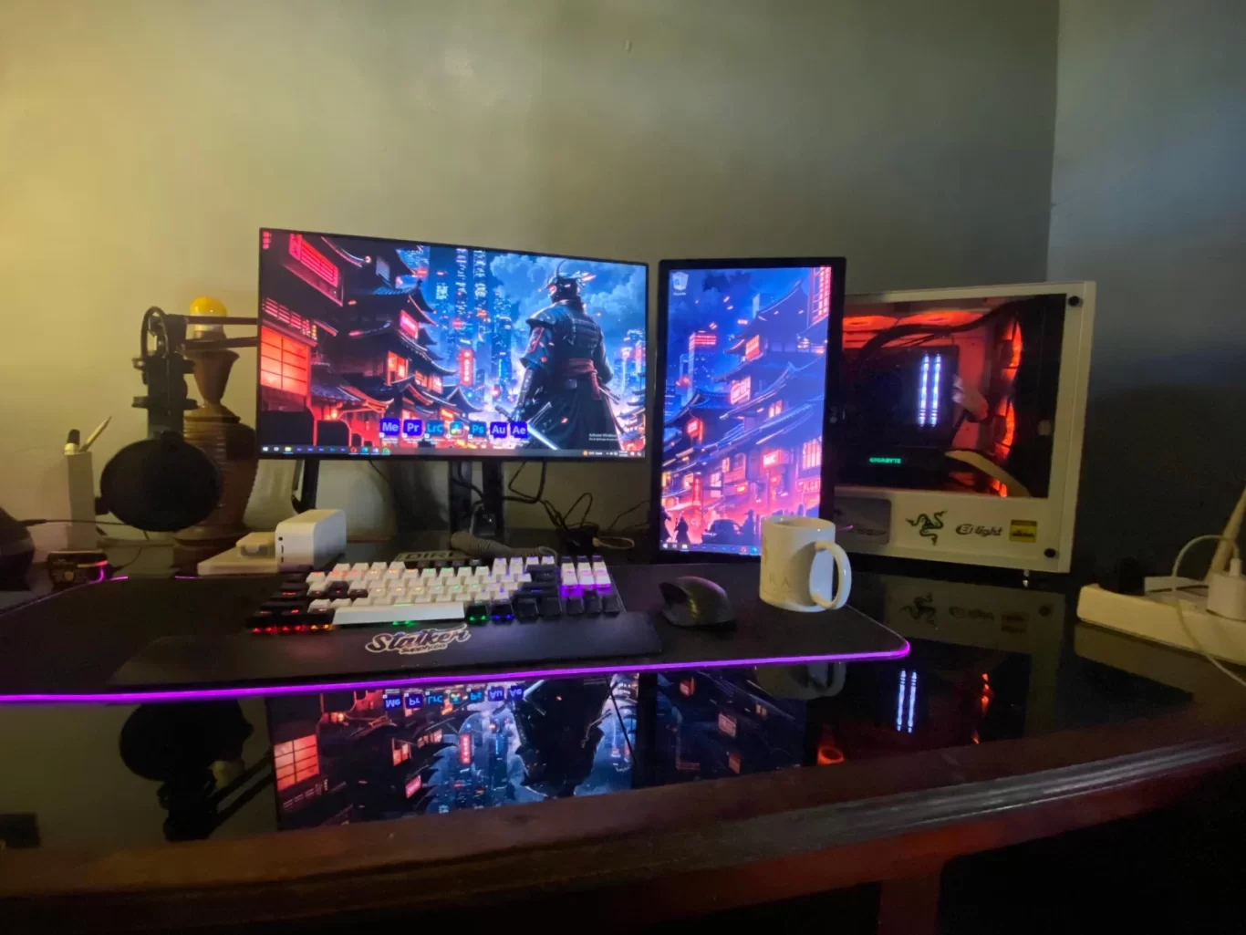 A dual monitor computer setup sits on a reflective dark desk, displaying vibrant blue and red cityscapes, with a white RGB keyboard and a gaming PC tower.