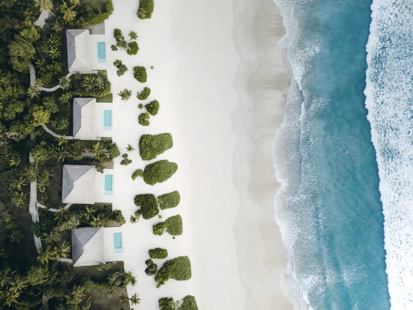 An aerial view shows white-roofed villas with small blue pools nestled in lush green foliage, bordering a pristine white sand beach and turquoise ocean waves.