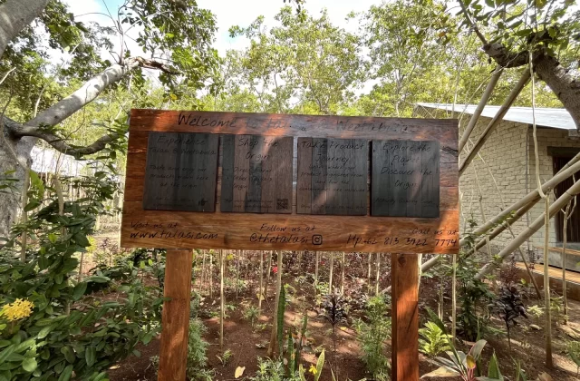 A wooden welcome sign for Talasi Weetabula stands in a vibrant garden with small plants and reddish soil, framed by lush green trees.