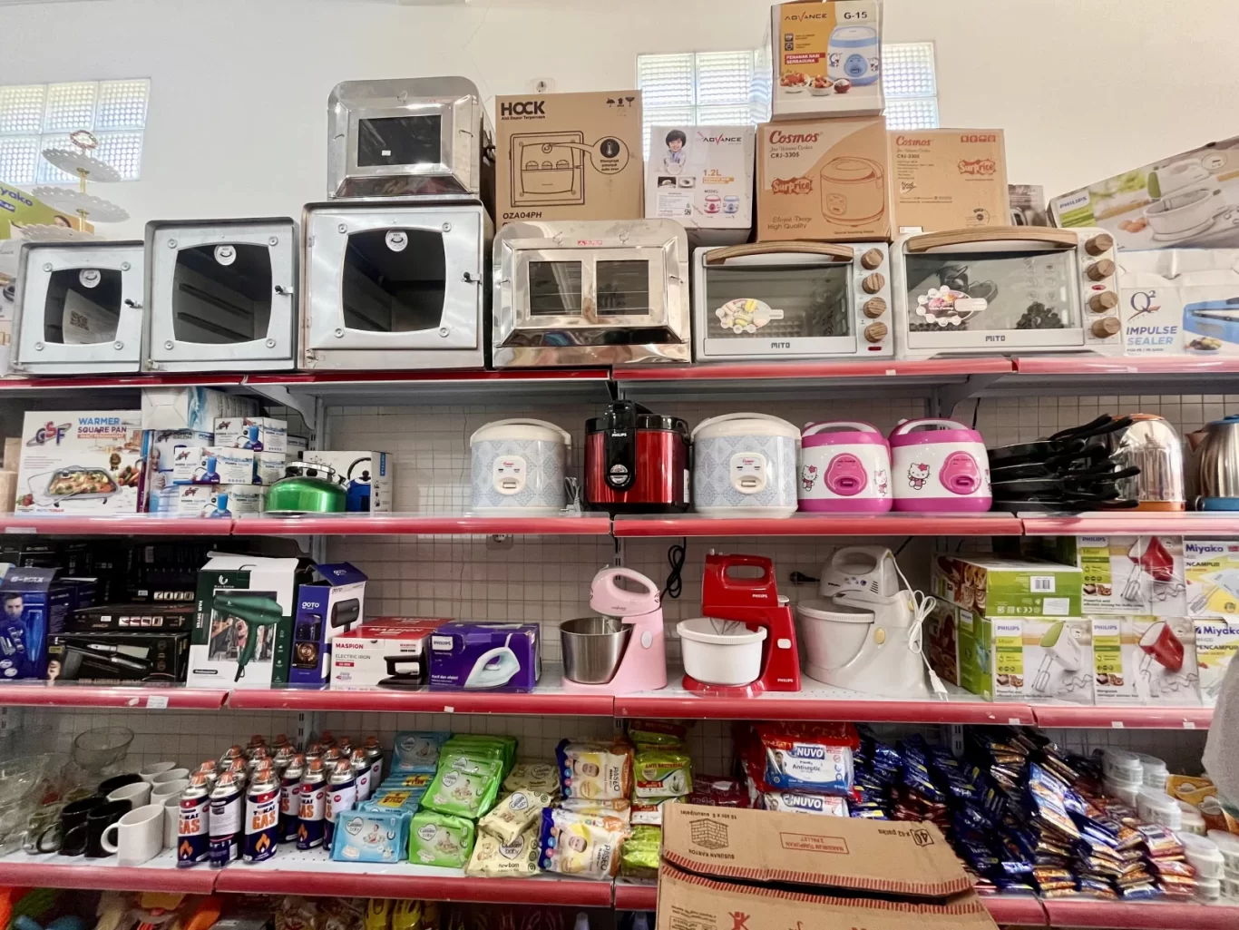 Brightly lit store shelves display a wide array of kitchen appliances, including silver ovens, colorful rice cookers, and stand mixers.