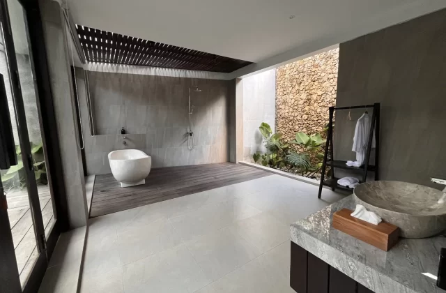 A modern villa bathroom features a white freestanding tub, grey tiled walls, a dark wood deck, and an open-air section with a stone wall and green plants.