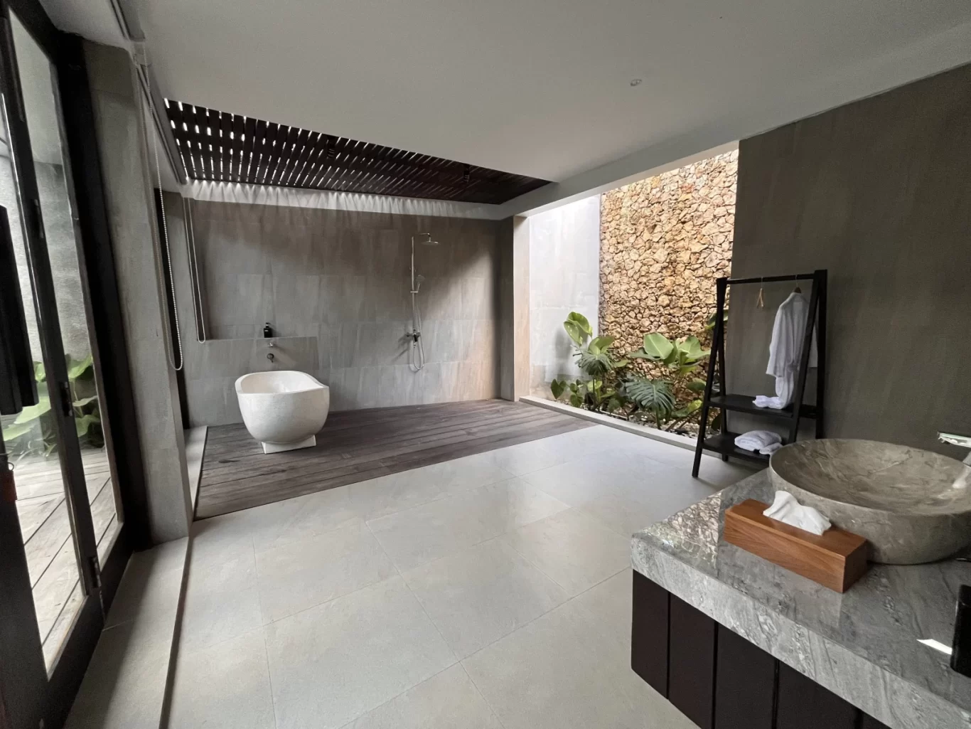 A modern villa bathroom features a white freestanding tub, grey tiled walls, a dark wood deck, and an open-air section with a stone wall and green plants.