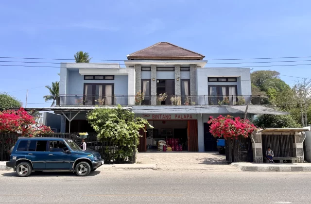A light blue two-story building with 'Toko Bintang Palapa' on its ground floor, a blue SUV, and pink bougainvillea line a sunny street.