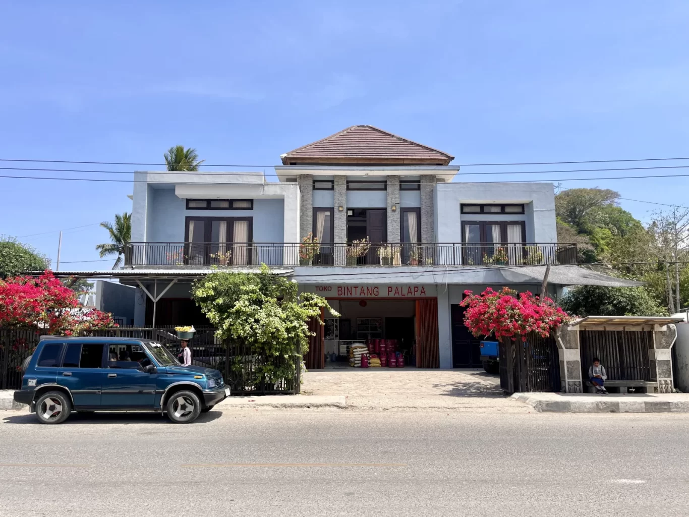 A light blue two-story building with 'Toko Bintang Palapa' on its ground floor, a blue SUV, and pink bougainvillea line a sunny street.