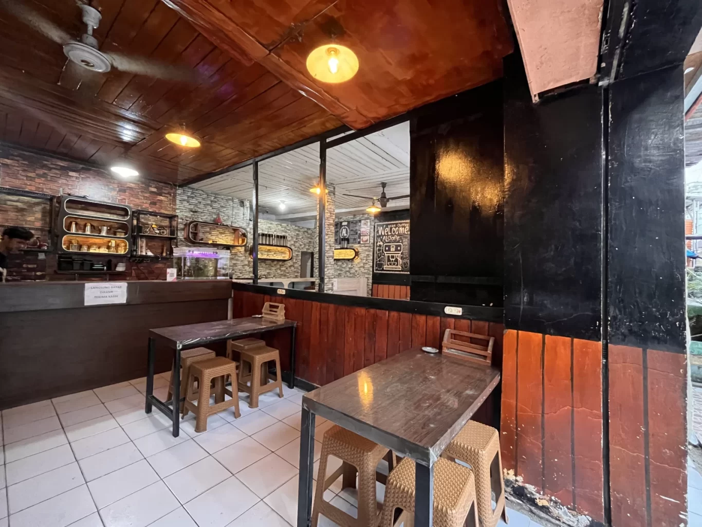A warmly lit cafe interior features a wooden ceiling, brick walls, and white tiled floor, with tables, stools, and a counter where a person stands.