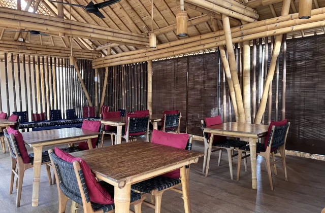 Interior view of a spacious restaurant with bamboo structure, thatched roof, wooden tables, and chairs with red cushions, alongside a blue lounge area.