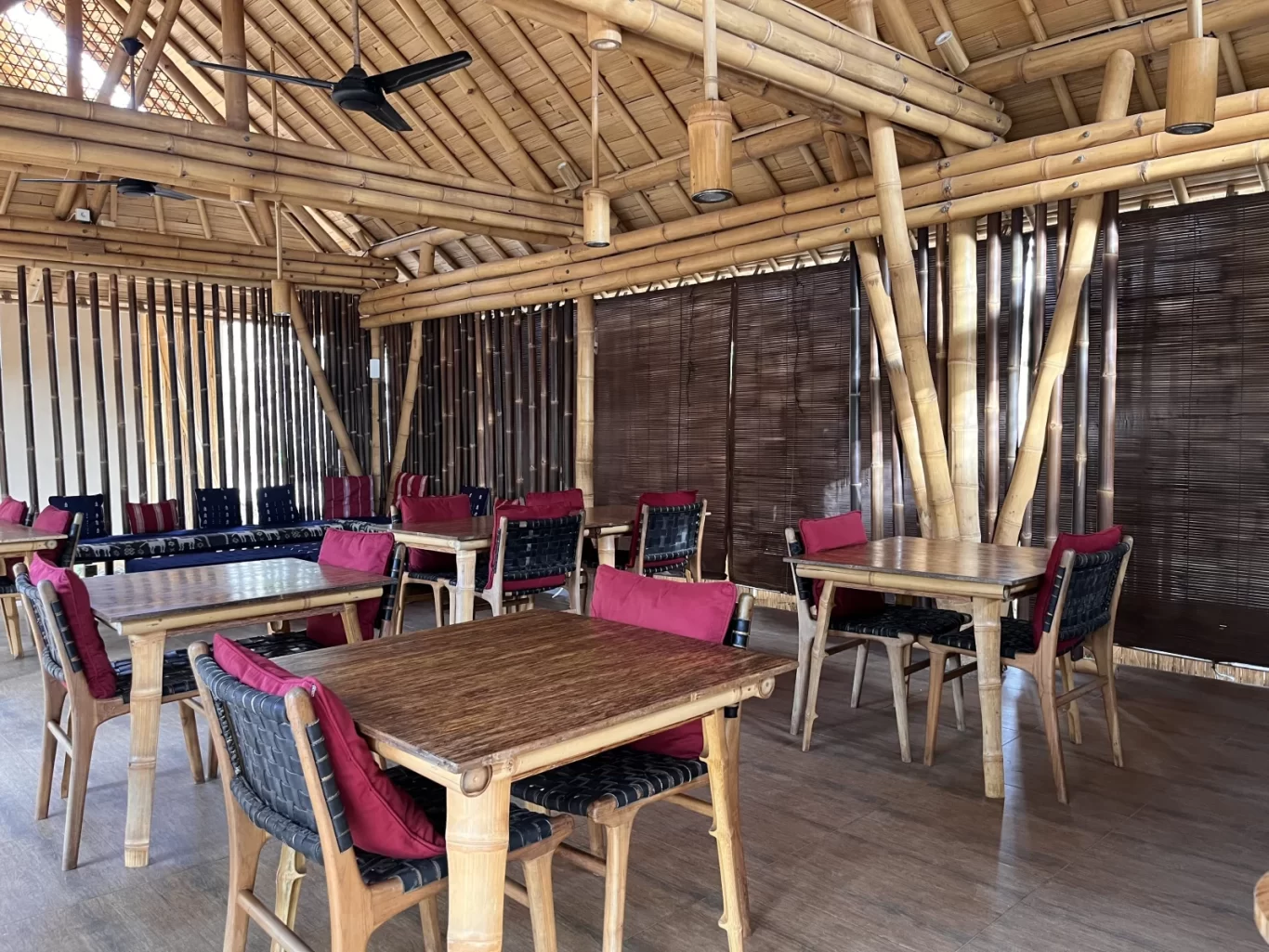 Interior view of a spacious restaurant with bamboo structure, thatched roof, wooden tables, and chairs with red cushions, alongside a blue lounge area.