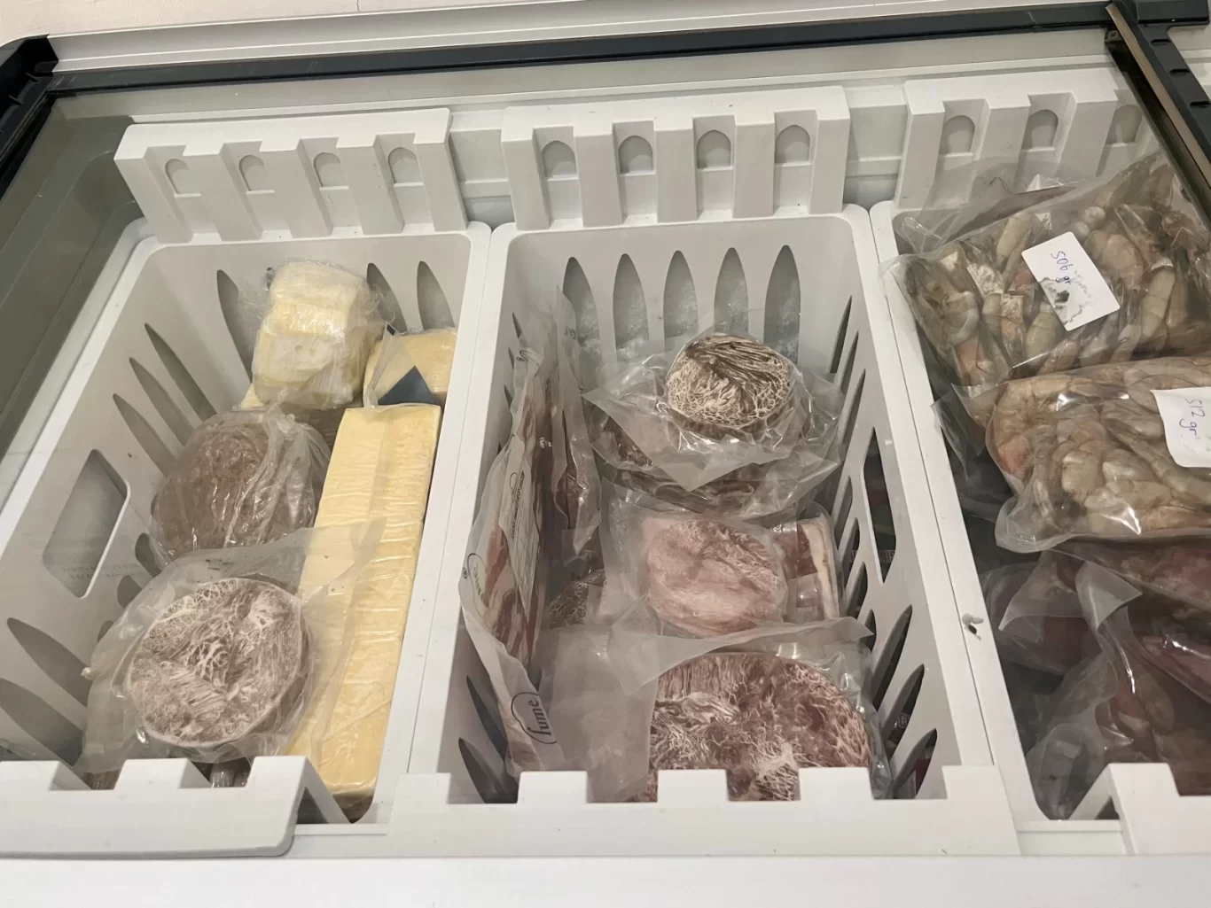 Visible inside a white freezer chest are various packaged frozen foods, including blocks of cheese, cuts of meat, and bags of shrimp.