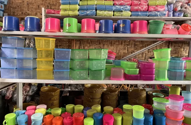 Shelves in a store are packed with a vibrant array of plastic household items, including cups, bowls, and storage containers, against a woven bamboo wall.