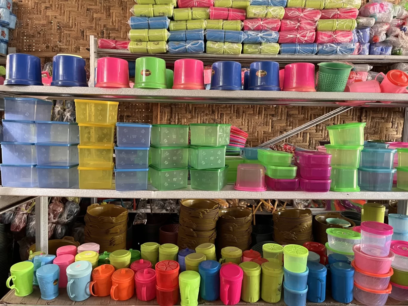 Shelves in a store are packed with a vibrant array of plastic household items, including cups, bowls, and storage containers, against a woven bamboo wall.
